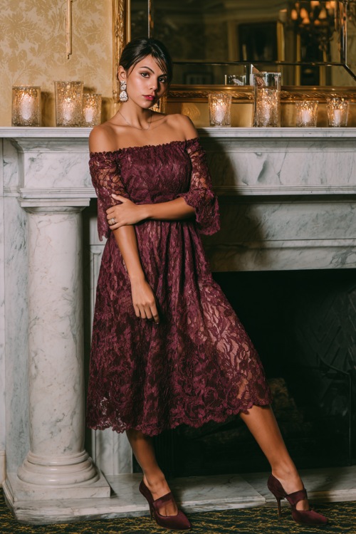 Full body of a woman in a wine-colored lace midi wedding guest dress with scalloped sleeves, pearl earrings, and kitten heels