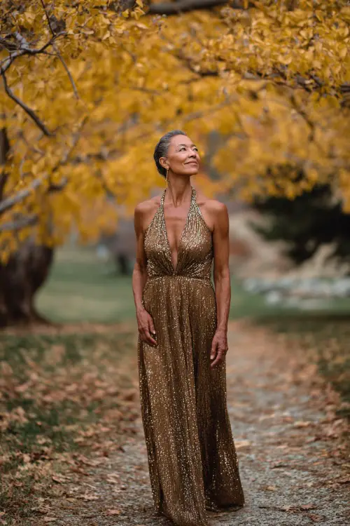 Full body of a woman over 50 in a bronze sequin-embellished gown with modest neckline and flowing skirt, standing beneath golden fall trees near a ceremony aisle