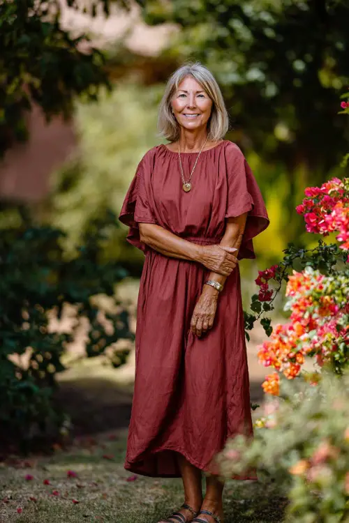 Full body of a woman over 50 in a light burgundy midi dress with short flutter sleeves and flowy hem, standing in a sunlit garden with soft fall florals