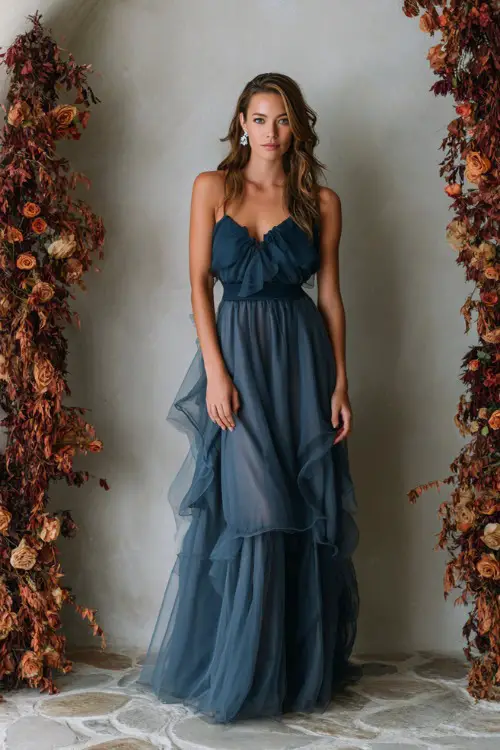 Full body of a woman over 50 in a slate blue tulle gown with layered skirt and sheer cape, posing beside a romantic wedding arch with cascading rust florals 