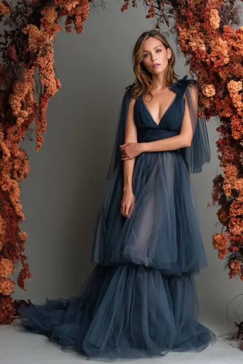 Full body of a woman over 50 in a slate blue tulle gown with layered skirt and sheer cape, posing beside a romantic wedding arch with cascading rust florals