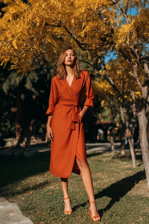 Full body of a woman wearing a burnt orange long-sleeve wrap dress with a midi hemline and block heels, standing under golden trees at an outdoor October wedding