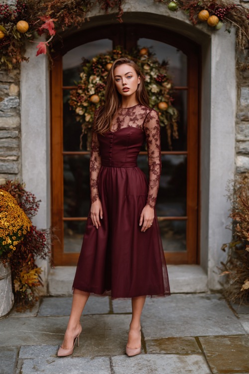 Full body of a woman wearing a maroon A-line midi wedding guest dress with sheer lace long sleeves and low heels, standing in front of a country estate with November wreaths and fall accents