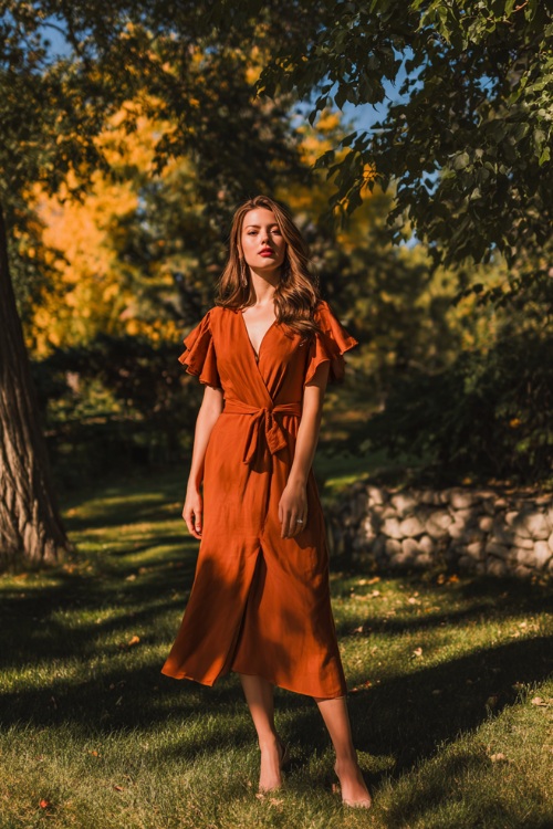 Full body of a woman wearing a rust-colored midi wedding guest dress with flutter sleeves and a tied waist, standing under early fall trees with warm sunlight at an outdoor wedding