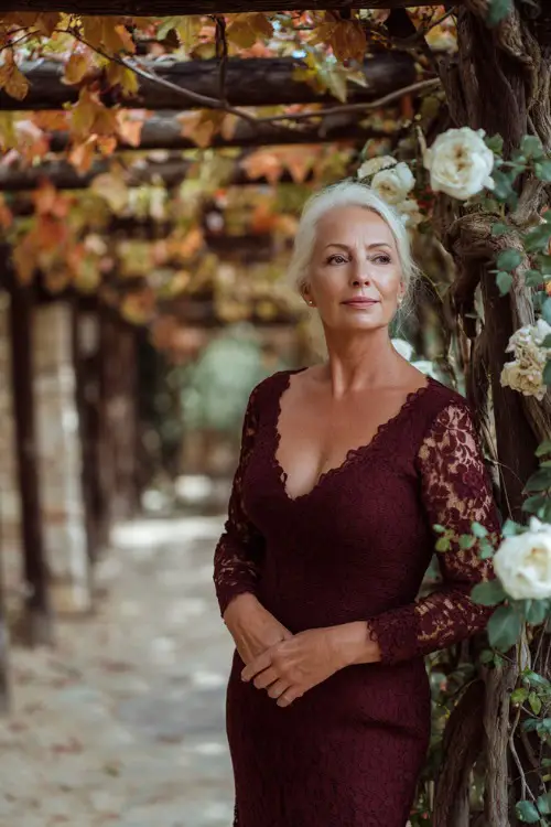 Mature woman in a long-sleeve maroon lace dress with slight mermaid hem, standing under a wooden pergola decorated with fall leaves and white roses 