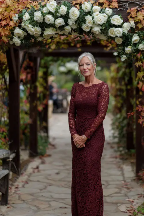 Mature woman in a long-sleeve maroon lace dress with slight mermaid hem, standing under a wooden pergola decorated with fall leaves and white roses