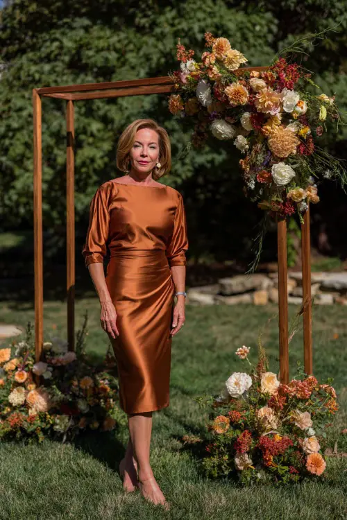 Mature woman wearing a copper satin midi dress with boat neckline and three-quarter sleeves, posing beside a wooden ceremony arch with early fall florals 