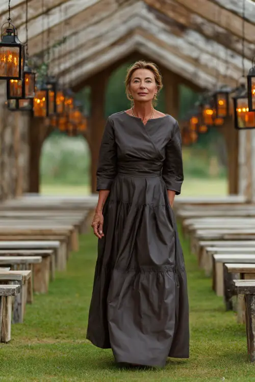 Mature woman wearing a warm charcoal wrap dress with three-quarter sleeves and a layered skirt, walking through a wedding aisle lined with rustic benches and hanging lanterns
