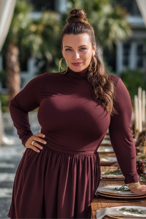 Plus-size woman in a burgundy mock-neck fit-and-flare dress with long sleeves and gold hoops, posing beside an outdoor reception table decorated with pinecones and candles 