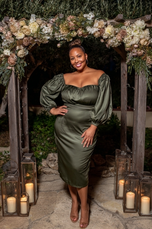 Plus-size woman in a dark olive green satin midi dress with a ruffle neckline and balloon sleeves, standing in front of a decorated arbor with neutral florals and lanterns
