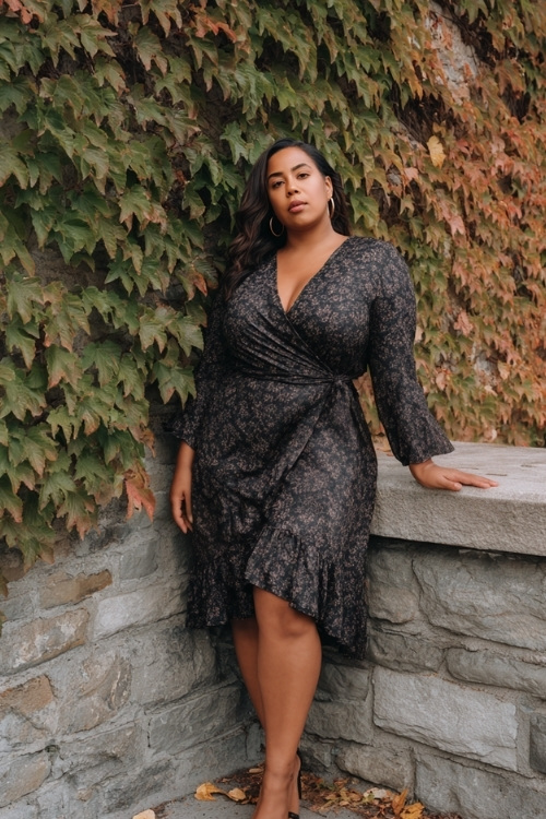 Stylish plus-size woman in a long-sleeve wrap dress with a dark floral print and ruffle hem, block heels and drop earrings, posing by a stone wall covered in fall ivy 