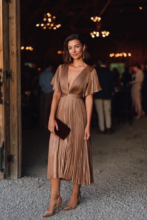 Woman in a mocha pleated chiffon midi dress with flutter sleeves and subtle shimmer, accessorized with a velvet clutch and strappy heels, standing inside a candlelit barn wedding venue 