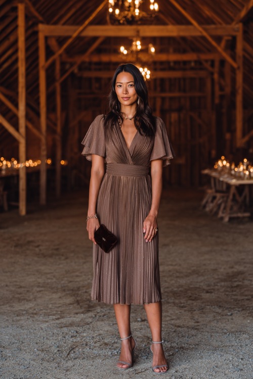 Woman in a mocha pleated chiffon midi dress with flutter sleeves and subtle shimmer, accessorized with a velvet clutch and strappy heels, standing inside a candlelit barn wedding venue 
