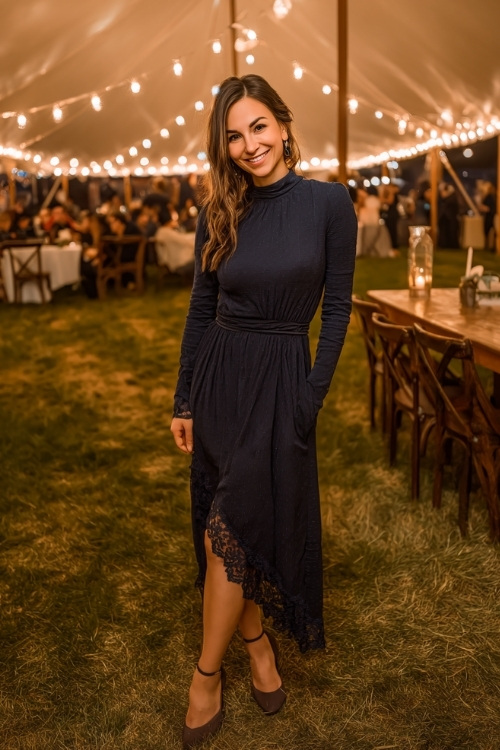 Woman in a navy long-sleeve wedding guest dress with lace trim, gathered waist, and low heels, posing under a string-light canopy at an October evening wedding 