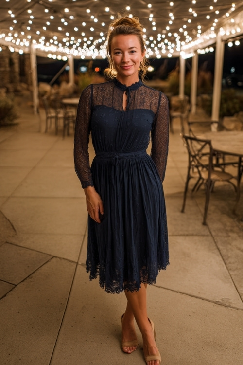 Woman in a navy long-sleeve wedding guest dress with lace trim, gathered waist, and low heels, posing under a string-light canopy at an October evening wedding 