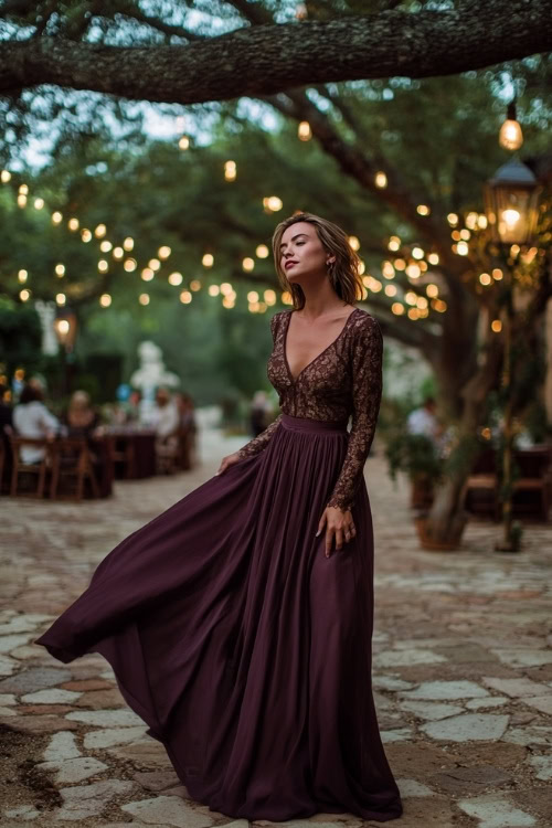 Woman in a plum-toned gown with lace long sleeves, fitted bodice, and flowing skirt, standing under a string-lit tree canopy at a romantic outdoor ceremony