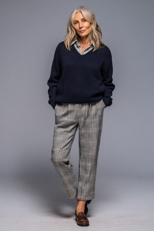 Woman in a simple navy pullover sweater layered with a button-up shirt underneath, gray plaid pants, and loafers, in a soft studio background 