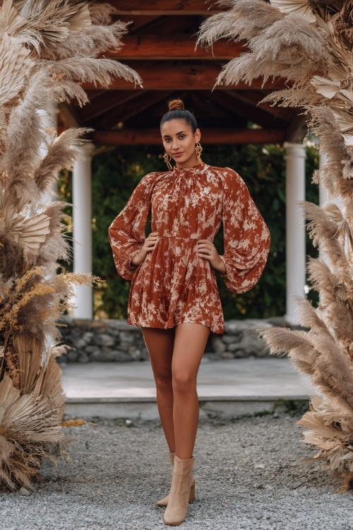 Woman in a terracotta floral print midi dress with long bell sleeves, gold jewelry, and ankle boots, posing near an outdoor wedding arch with dried pampas grass and leaves 