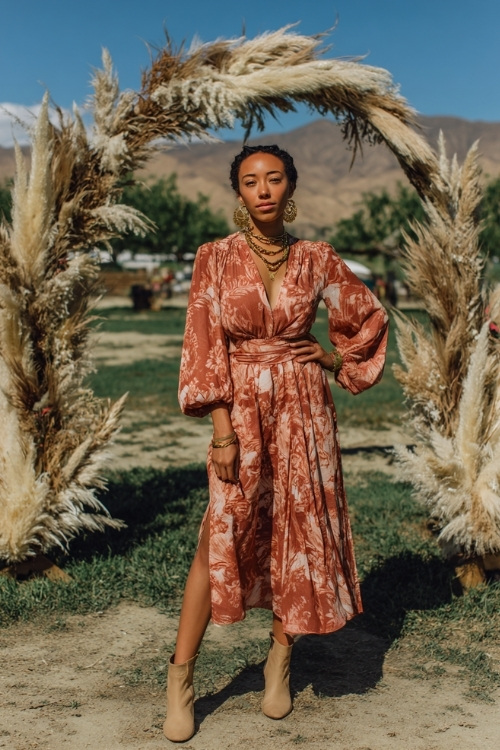 Woman in a terracotta floral print midi dress with long bell sleeves, gold jewelry, and ankle boots, posing near an outdoor wedding arch with dried pampas grass and leaves