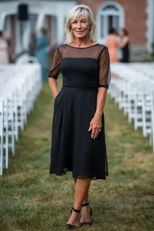 Woman over 50 in a classic black midi dress with short sheer sleeves and A-line silhouette, posing by white chairs lined up for an outdoor wedding 