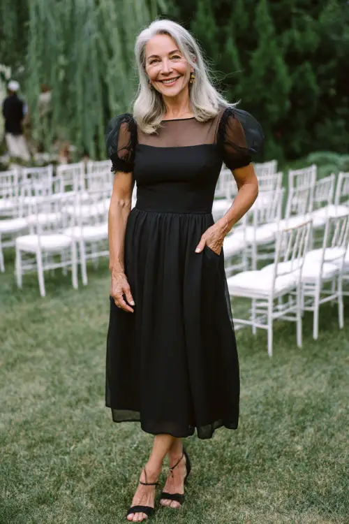 Woman over 50 in a classic black midi dress with short sheer sleeves and A-line silhouette, posing by white chairs lined up for an outdoor wedding