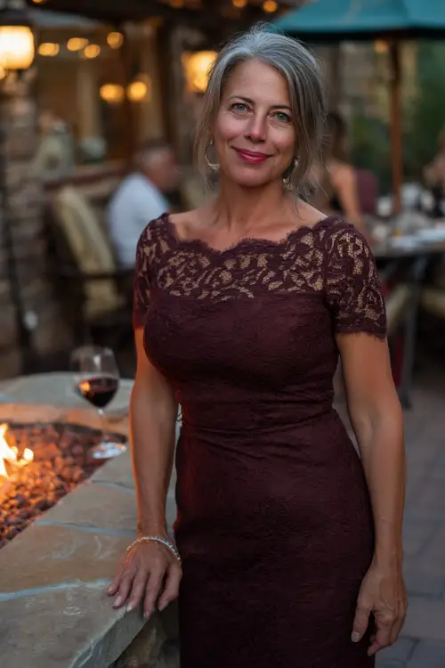 Woman over 50 in a dark burgundy dress with classic pencil silhouette and lace shoulders, posing near a fire-lit patio at an upscale outdoor wedding 