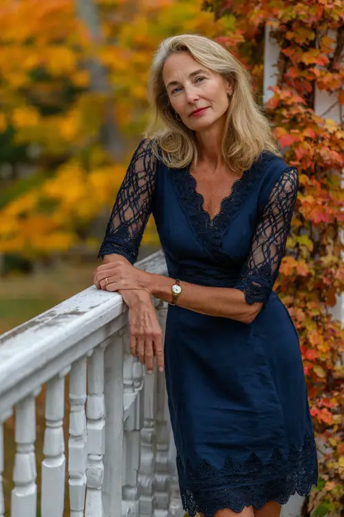 Woman over 50 in a navy blue knee-length shift dress with lace sleeves and scalloped hem, posing near a white fence wrapped in fall foliage