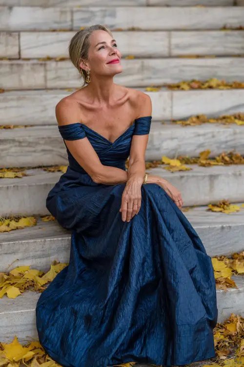 Woman over 50 in a navy blue off-shoulder floor-length gown with structured pleats and subtle shimmer, posing on marble steps surrounded by golden fall leaves 