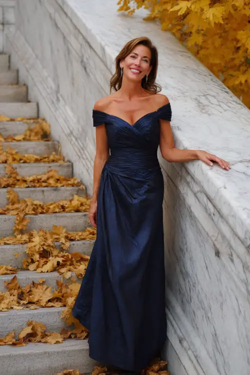 Woman over 50 in a navy blue off-shoulder floor-length gown with structured pleats and subtle shimmer, posing on marble steps surrounded by golden fall leaves