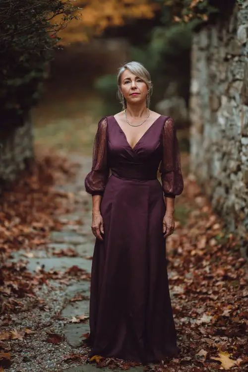 Woman over 50 in a plum fit-and-flare gown with sheer bishop sleeves and a subtle shimmer, standing on a stone path surrounded by crisp October leaves
