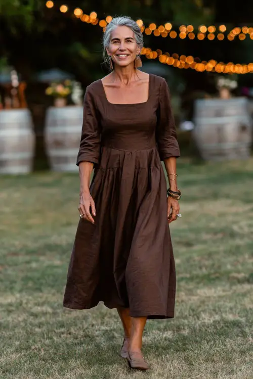 Woman over 50 in a rich mocha-colored midi dress with flared skirt and square neckline, walking across an open lawn with string lights and rustic barrels 