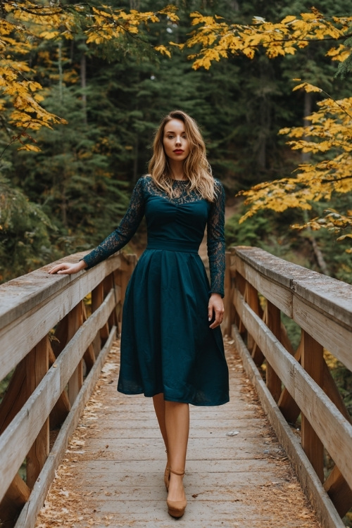 Woman wearing a dark teal A-line midi dress with long sleeves, lace neckline, and suede block heels, standing on a wooden bridge covered in golden leaves at a cozy October wedding in the forest 