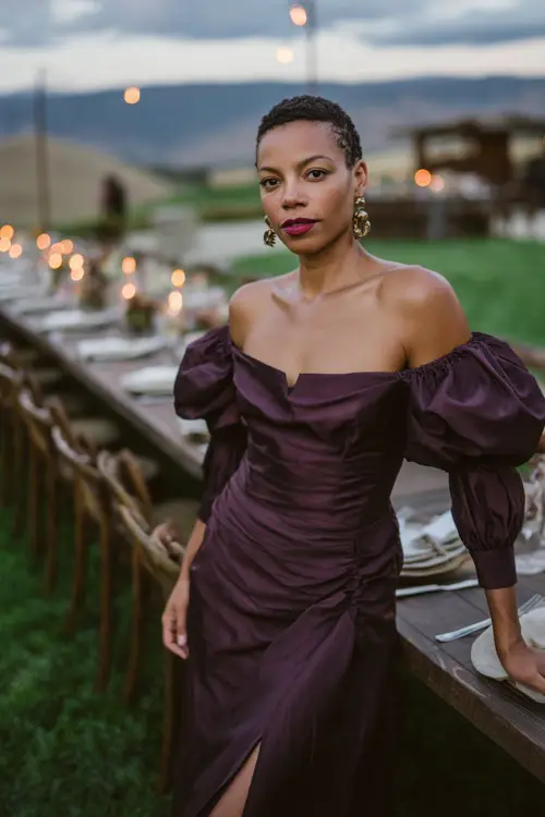 A woman in a dark plum satin midi dress with puff sleeves and slit, accessorized with metallic earrings, posing at an outdoor country wedding reception with candlelit tables 