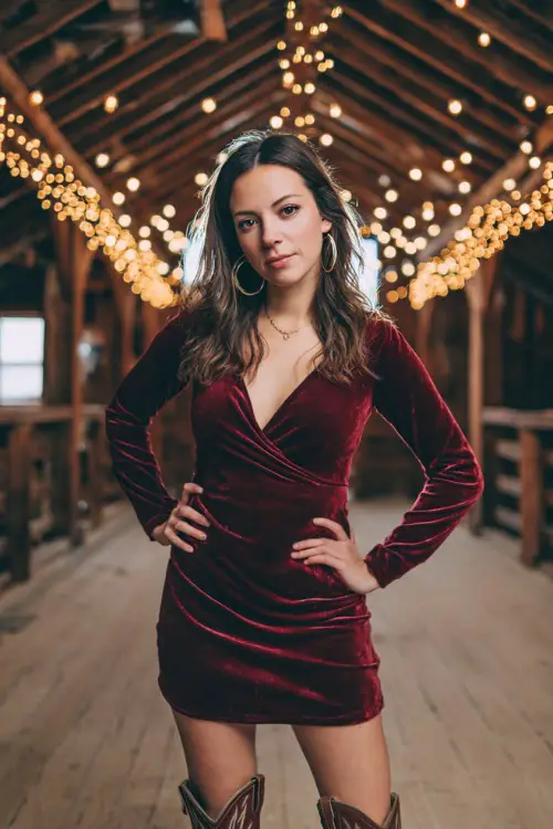 A woman in a maroon velvet skater dress with long sleeves, accessorized with cowboy boots and hoop earrings, standing inside a rustic barn lit with fairy lights 