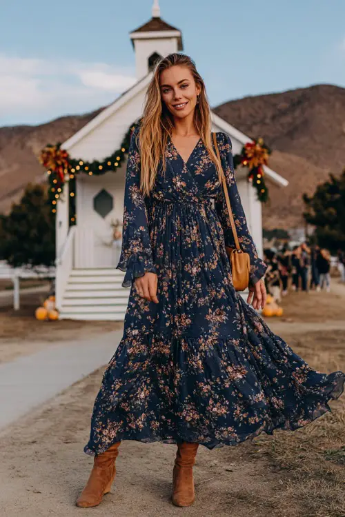 A woman in a navy floral print maxi dress with long sleeves and a flowy skirt, paired with suede boots and a crossbody clutch, posing in front of a countryside chapel with fall garlands