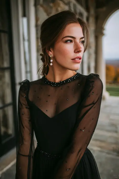 A woman wears a black chiffon gown with long sheer sleeves, beaded bodice, and pearl earrings in an elegant autumn-themed wedding venue