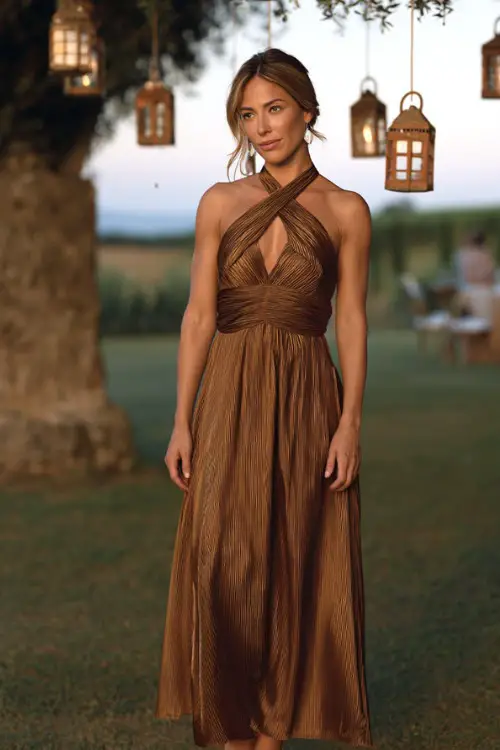 A woman wears a bronze pleated maxi dress with a halter neckline, styled with strappy block heels, standing at a countryside outdoor reception under hanging lanterns