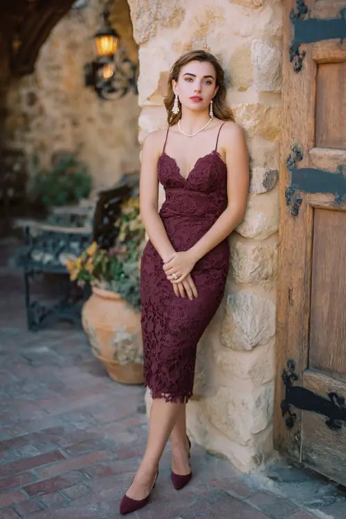 A woman wears a burgundy lace scalloped-hem cocktail dress, pumps, and pearl drop earrings at a rustic stone courtyard wedding 