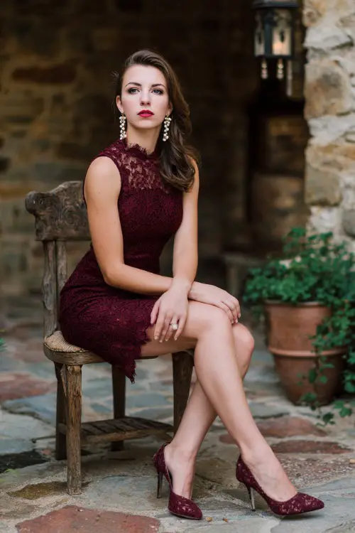 A woman wears a burgundy lace scalloped-hem cocktail dress, pumps, and pearl drop earrings at a rustic stone courtyard wedding