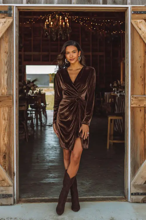A woman wears a chocolate brown velvet wrap dress with knee-length hem, paired with heeled boots and hoop earrings, standing at the entrance of a rustic wooden barn wedding 