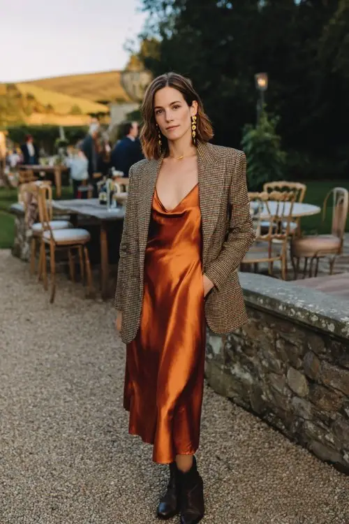 A woman wears a copper satin slip dress layered with a plaid wool blazer, paired with ankle boots and gold earrings