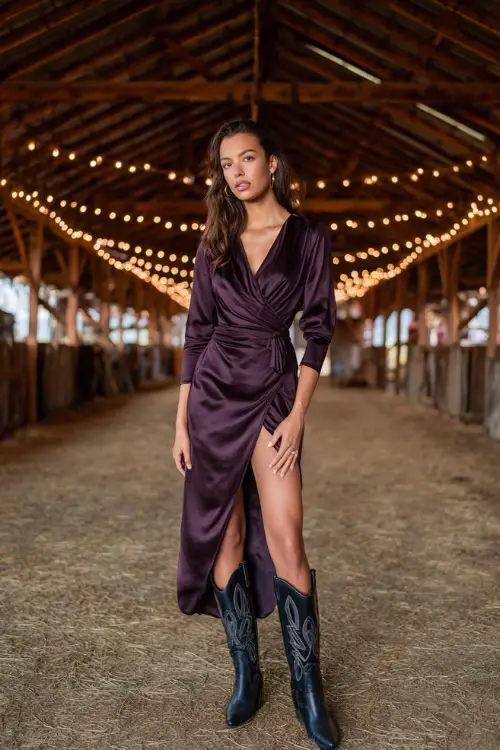 A woman wears a dark plum satin wrap dress with slit, styled with black leather cowboy boots, standing inside a rustic barn venue glowing with fairy lights 