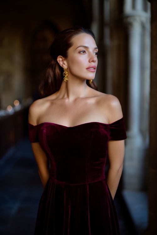 A woman wears a deep burgundy velvet off-the-shoulder gown with a fitted bodice, floor-length skirt, and gold statement earrings at a candlelit cathedral wedding