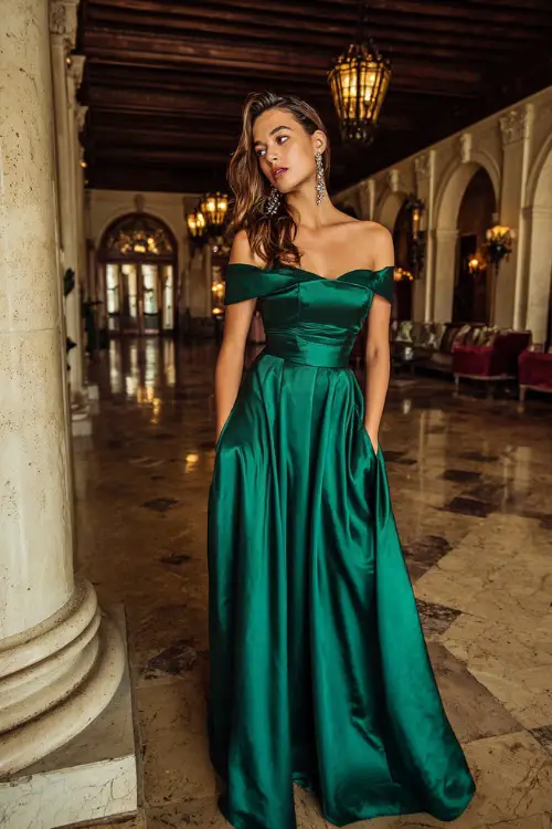 A woman wears a deep emerald green satin gown with an off-the-shoulder neckline, floor-length skirt, and silver drop earrings in a grand ballroom setting 