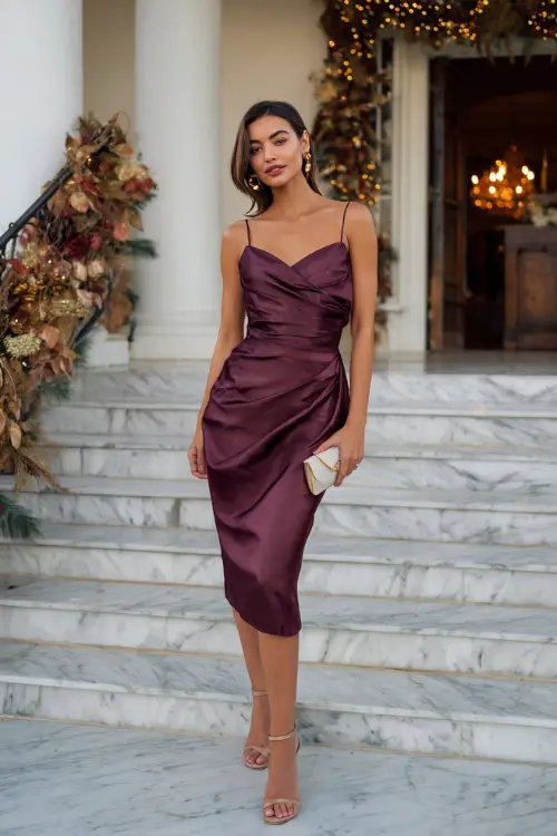 A woman wears a deep plum satin midi dress with a draped neckline and slit, styled with strappy heels and a clutch