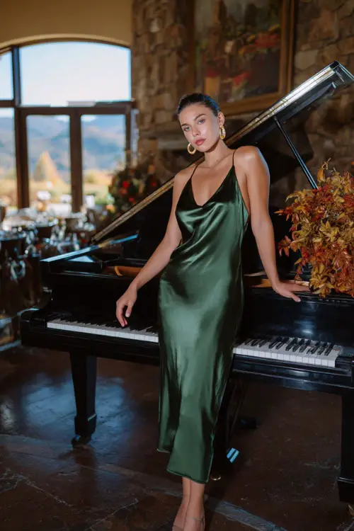 A woman wears a forest green satin slip dress with a cowl neckline, styled with gold sandals and earrings