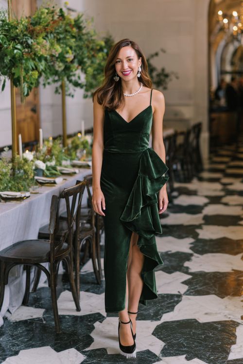 A woman wears a forest green velvet gown with asymmetrical ruffle details, pearl accessories, and sleek black heels