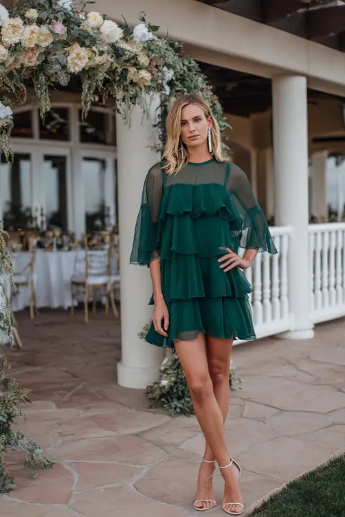 A woman wears a hunter green chiffon cocktail dress with layered ruffles and sheer sleeves, styled with silver earrings and strappy heels 
