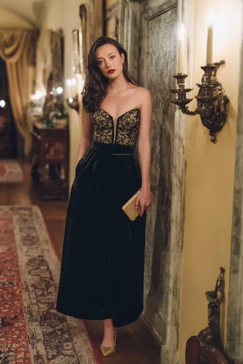 A woman wears a midnight black velvet A-line gown with an embroidered bodice, gold heels, and a matching clutch in a candlelit estate venue 