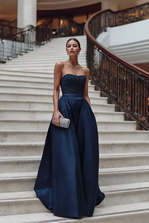 A woman wears a midnight blue A-line gown with embroidered bodice, silver clutch, and diamond studs at a grand indoor staircase setting 